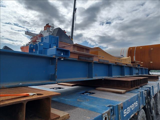 Metal beams and industrial equipment on transport platforms, with a ship docked in the background.