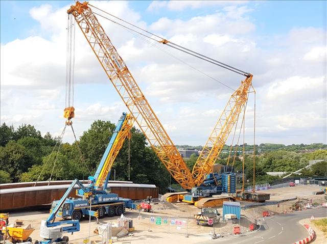 Two massive cranes assembling heavy equipment at a busy construction site, highlighting their scale and precision.