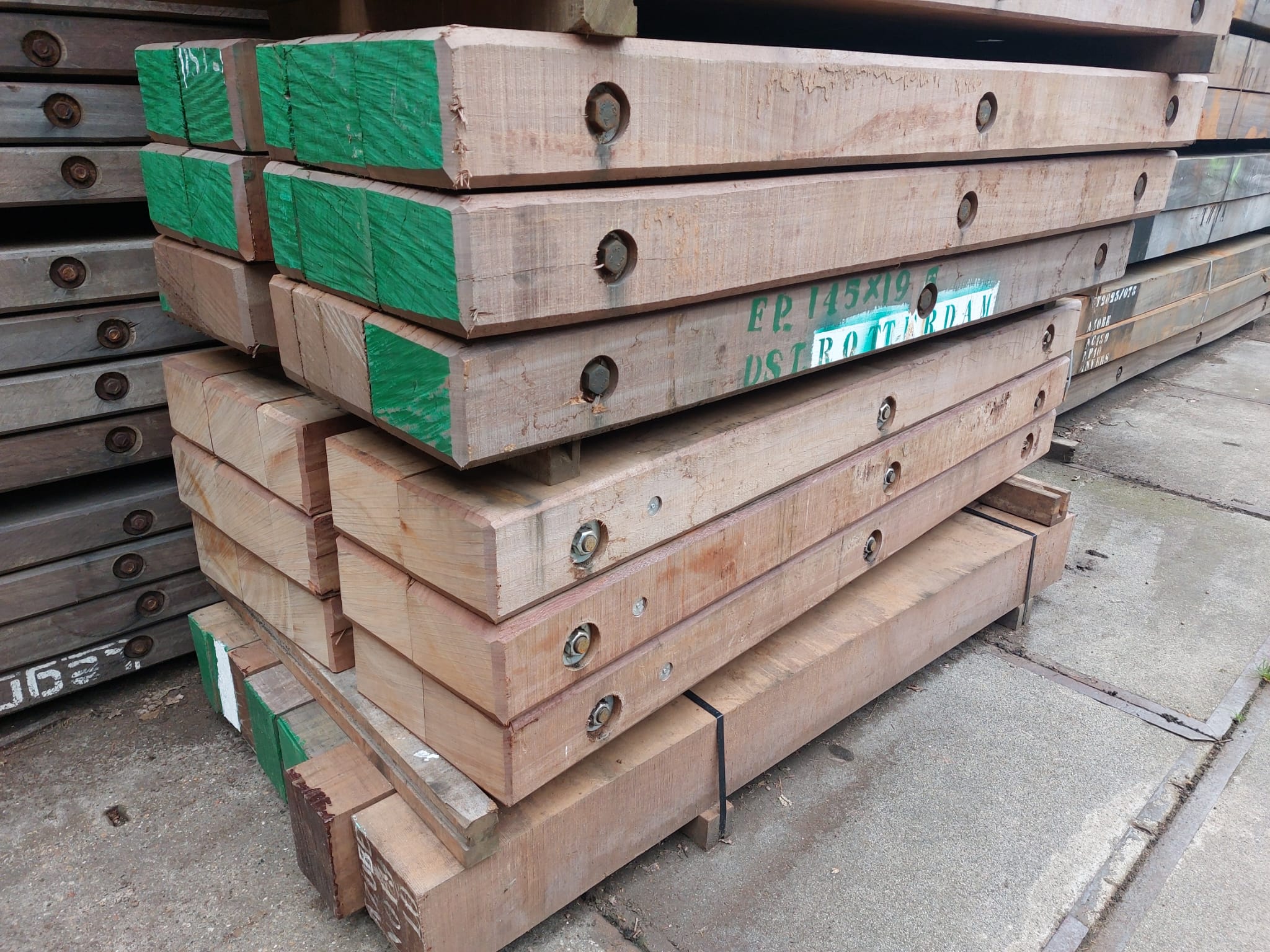 Stack of timber mats with green-painted ends, showing size or type markings, stored outdoors